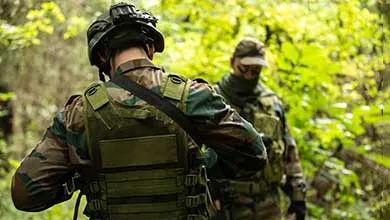 Rear view of a soldier in camouflage, tactical vest, and helmet, standing in a forest with another soldier partially visible