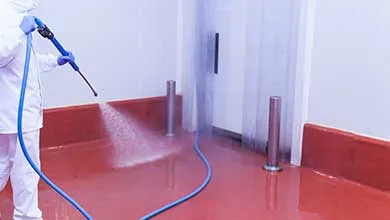 Person in white protective suit and blue gloves pressure washing a red industrial floor.