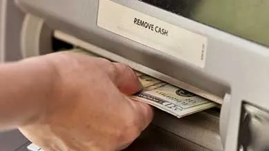 Hand withdrawing twenty-dollar bills from an ATM cash dispenser labeled "REMOVE CASH."