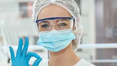 A healthcare worker wearing a hairnet, goggles, face mask, and gloves makes an "OK" gesture with her hand.