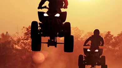 Two people on ATVs are silhouetted against an orange sunset; one is airborne in mid-jump.