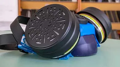 Close-up of a blue respirator mask with two large black circular filters on each side, resting on a green table.