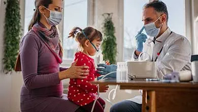 A doctor helps a young girl use a nebulizer while she sits on her mother's lap.
