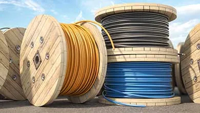 Low-angle view of large wooden spools of industrial cable outdoors. An orange cable spool is in the foreground, beside a stack with black cable on top and blue cable below.