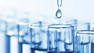 A row of clear glass test tubes on a surface with a dropper holding a single liquid droplet above them, against a bright, softly blurred background.