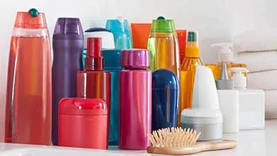 Colorful bottles and jars of toiletries arranged on a white surface, with a wooden hairbrush in front and a stack of white towels behind.