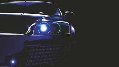 Close-up of a dark car with bright blue headlights shining in the shadows against a black background.
