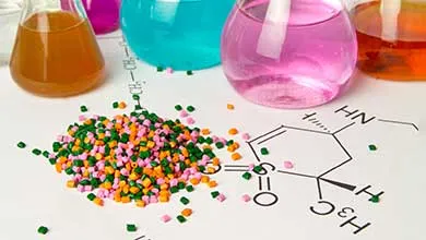 Small green, orange, and pink plastic pellets rest on a surface with chemical structure drawings. Behind them, glass flasks hold orange, blue, and pink liquids.
