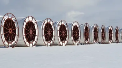 A row of large silver corrugated spools with red spoked ends sits on snow under a cloudy sky.